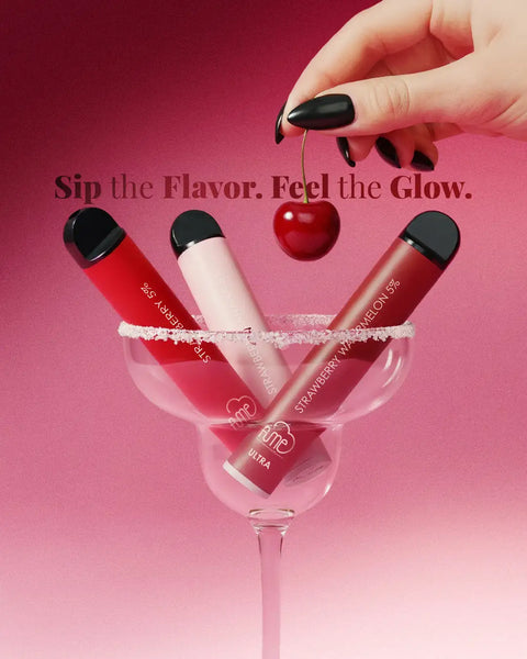 Three sleek, cylindrical disposable vape pens in vibrant red and soft pink hues with glossy finishes are nestled inside a clear martini glass rimmed with sugar.